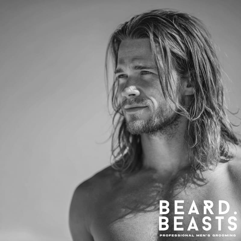 Portrait of a male model with long, straight blonde hair, styled in a relaxed, natural surfer look against a clear sky.