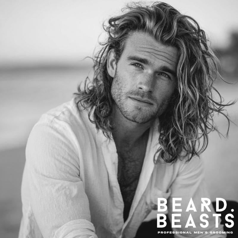 Portrait of a male model with long, wavy, textured hair, styled in a relaxed surfer look, in a black and white beach setting.