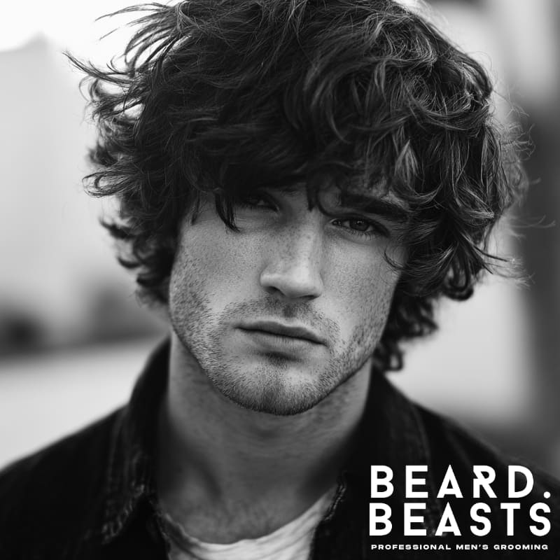 Male model with a textured hairstyle, showcasing soft waves and volume, set against a blurred city background for a candid, street-style look.