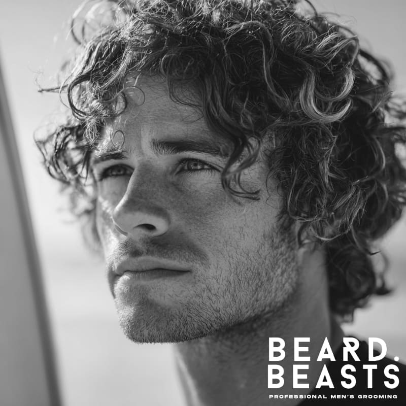Close-up of a male model with short, defined curly hair, showcasing a natural and textured surfer look under sunlight.