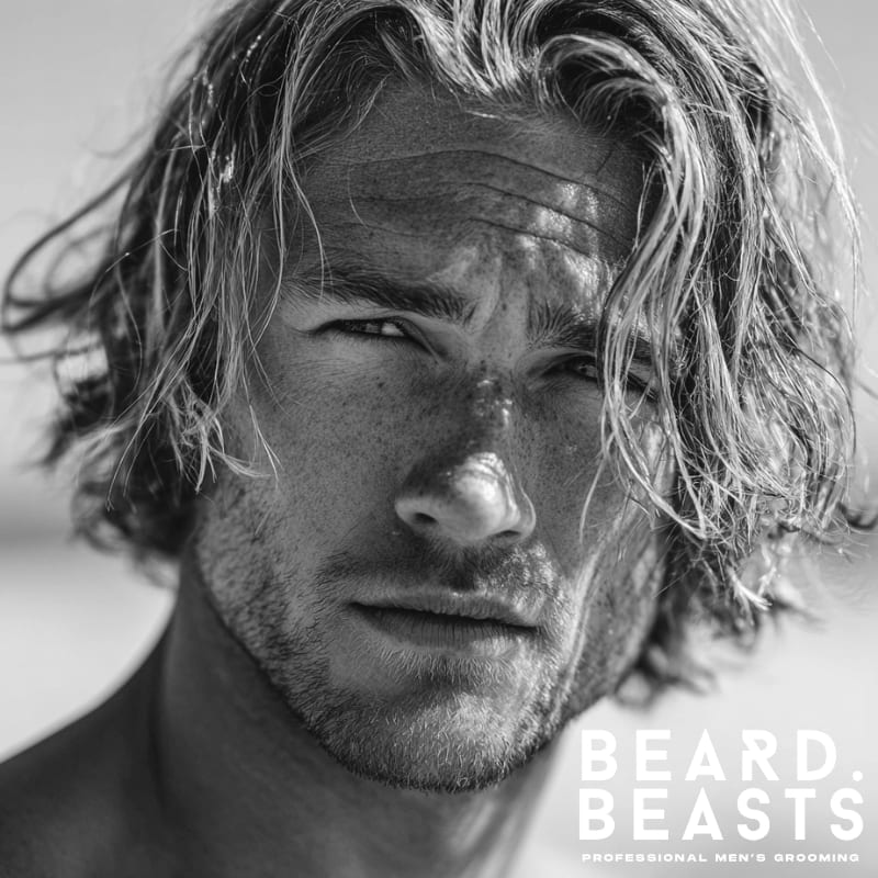 Close-up of a male model with natural, textured blonde surfer curtains, featuring long front layers parted softly to frame the face in a relaxed beach style.