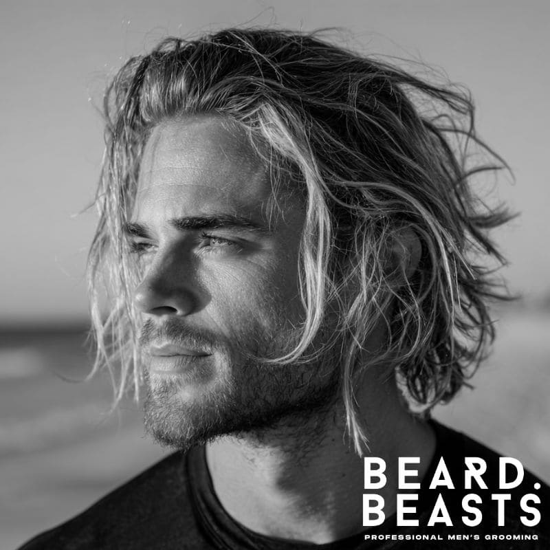 andsome male model with long, blonde, textured brushed-back surfer hair on a beach at sunset, showcasing a natural and effortless beach hairstyle.