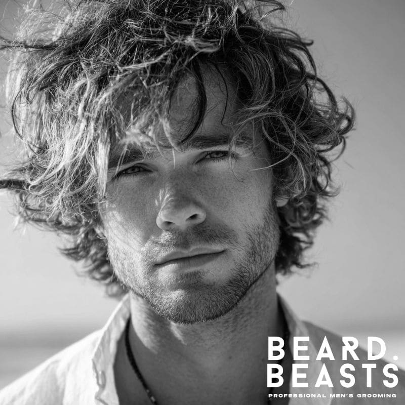 Close-up of a male model with a tousled shag haircut, featuring messy, natural blonde curls and a beachy, windswept look.