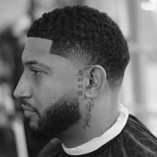 A fresh mid taper fade haircut, featuring a precise fade that blends seamlessly into a textured curly top. The sharp lineup and well-groomed beard enhance the clean and stylish look, making this fade haircut a great choice for those who want a polished yet modern appearance. The tattoo details on the side add a unique personal touch to the overall aesthetic.