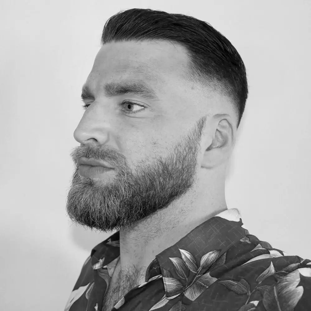 A three-quarter profile shot of a man with a short slick back haircut and a skin fade. He has a full beard and is wearing a red floral-patterned collared shirt.