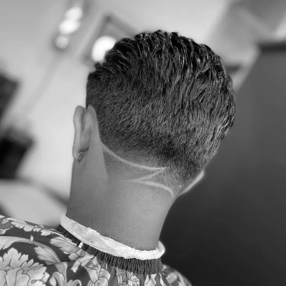 A view of the back of a person's head, showcasing a fresh haircut. The style features a low taper fade at the nape with a sharp, geometric line design shaved into it. The longer hair on top is styled in a textured slick back.