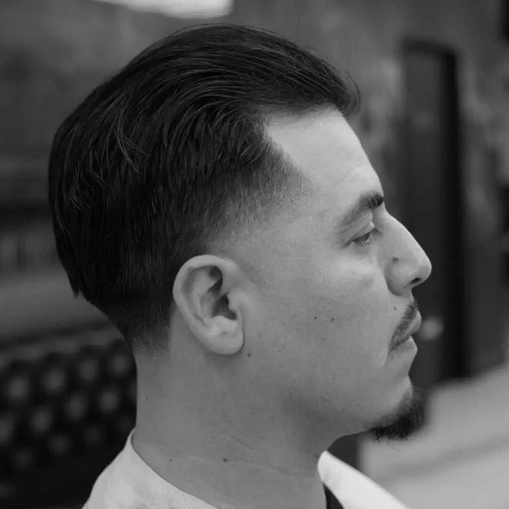 A profile view of a man showcasing a slick back haircut with a high taper fade. The longer hair on top is combed back neatly, while the sides are faded down cleanly from the temple. He has a goatee, and the photo is taken in a barber shop.