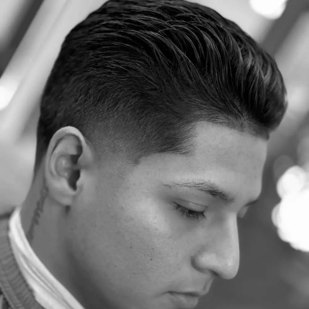 A close-up three-quarter profile of a man showcasing a slick back haircut with a mid taper fade. The voluminous hair on top is neatly combed back, while the side shows a clean taper starting in the middle of the head. The hairline is sharply defined.