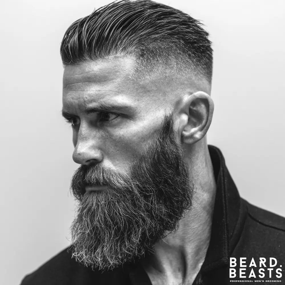 A close-up, black and white profile portrait of a man with a slick back skin fade haircut. His hair is neatly combed back on top, contrasting with sides that are faded down to the skin. He has a long, full, well-groomed beard and a serious expression.
