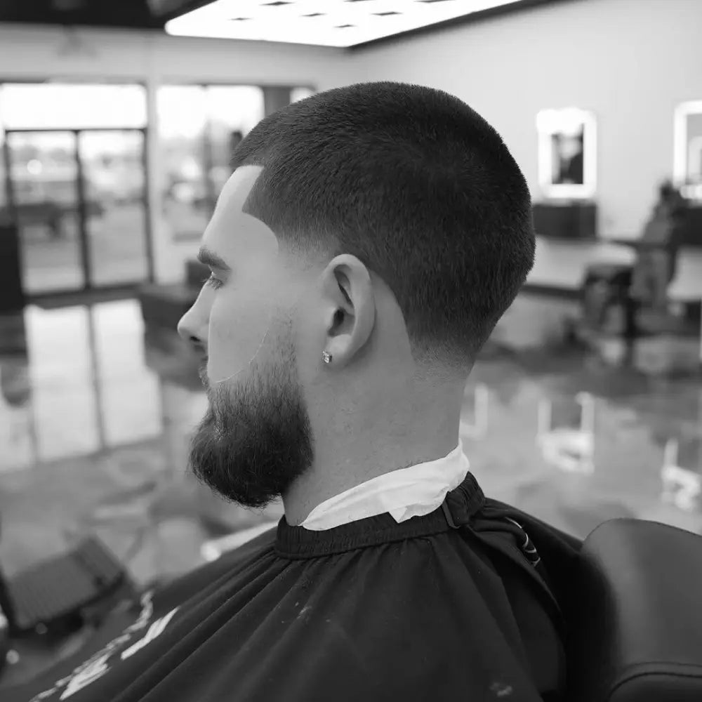 Man with a buzz cut and taper fade, featuring short, even hair on top with a gradual fade into the neckline and a neatly shaped beard for a clean, masculine look.
