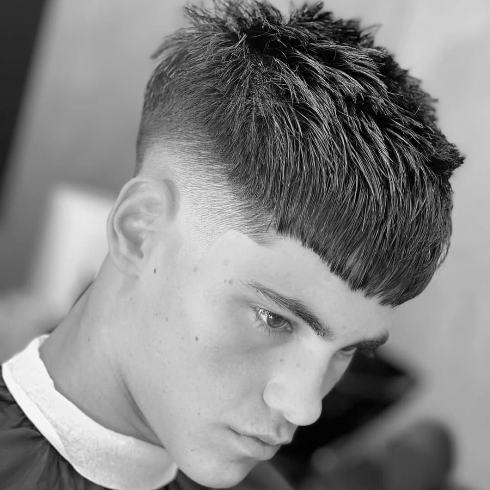 Young man with a mid-fade French crop hairstyle. The haircut features a clean and sharp mid-fade on the sides, seamlessly blending into the textured, choppy layers on top. The front showcases a straight, blunt fringe that adds a bold and modern edge to the overall look.