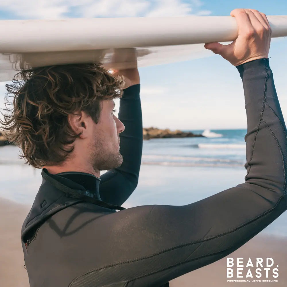 Man with textured wavy hair holding a surfboard at the beach, showing the natural volume and finish achieved with sea salt spray.