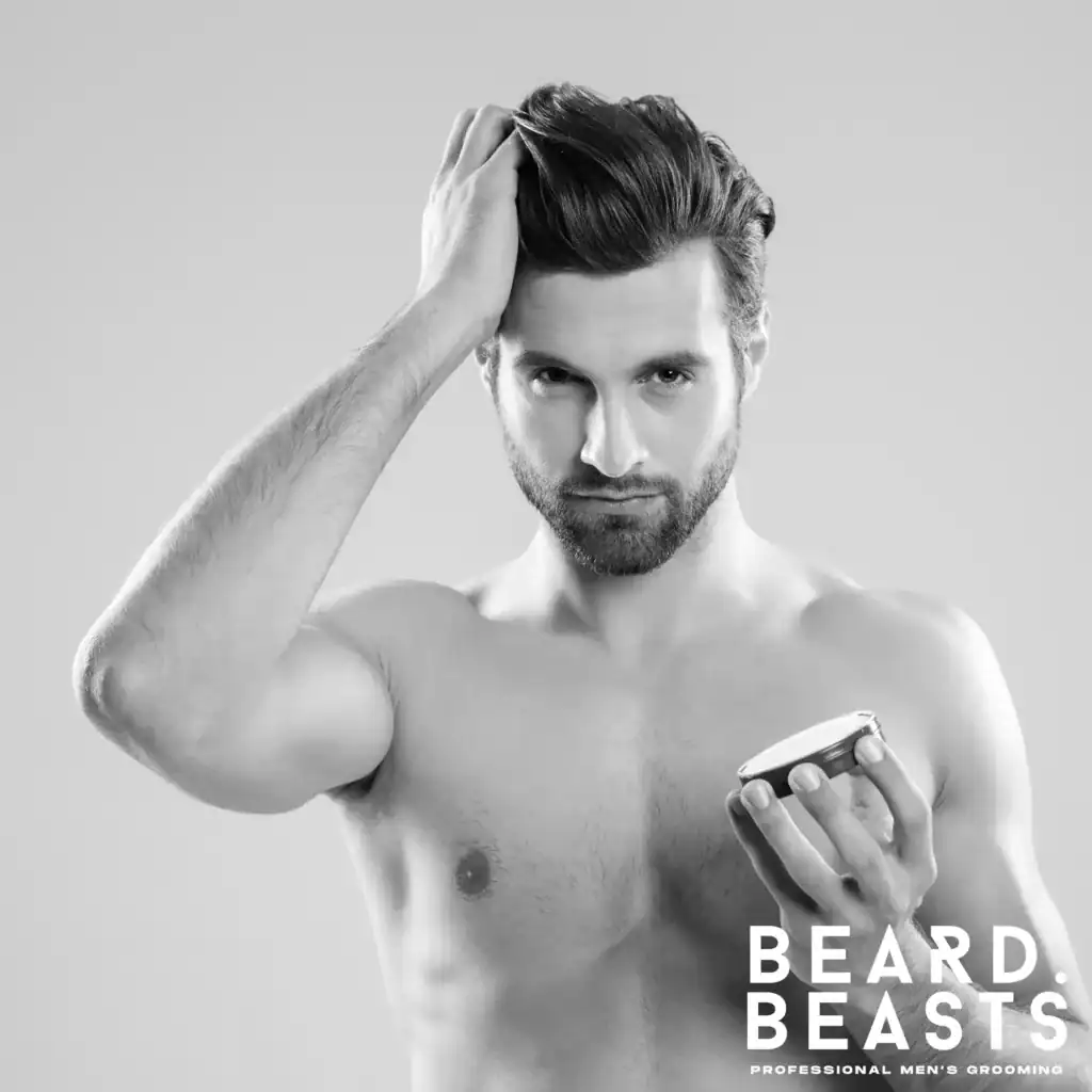 Shirtless man applying hair clay to style his textured hairstyle, holding a blue container in his hand.