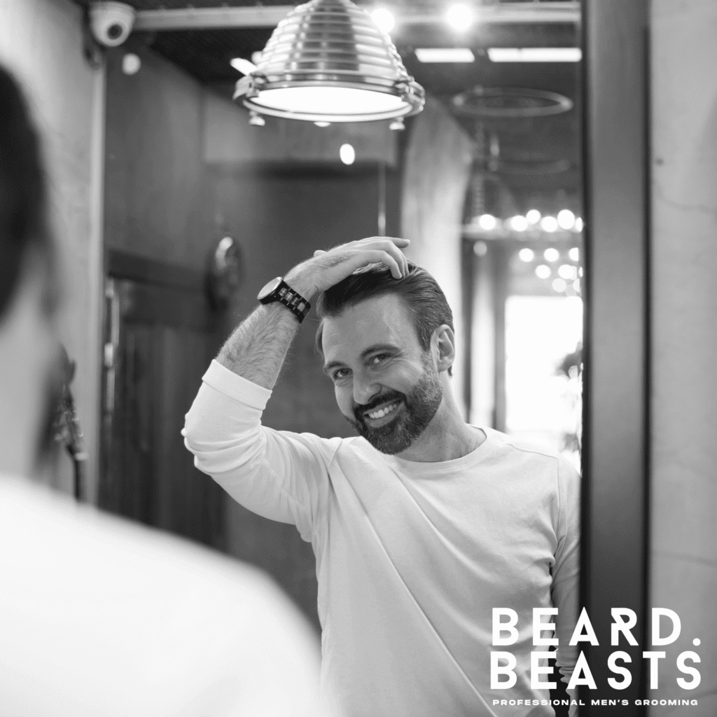 Smiling man styling his hair with matte clay in front of a mirror at a barbershop, wearing a white shirt.