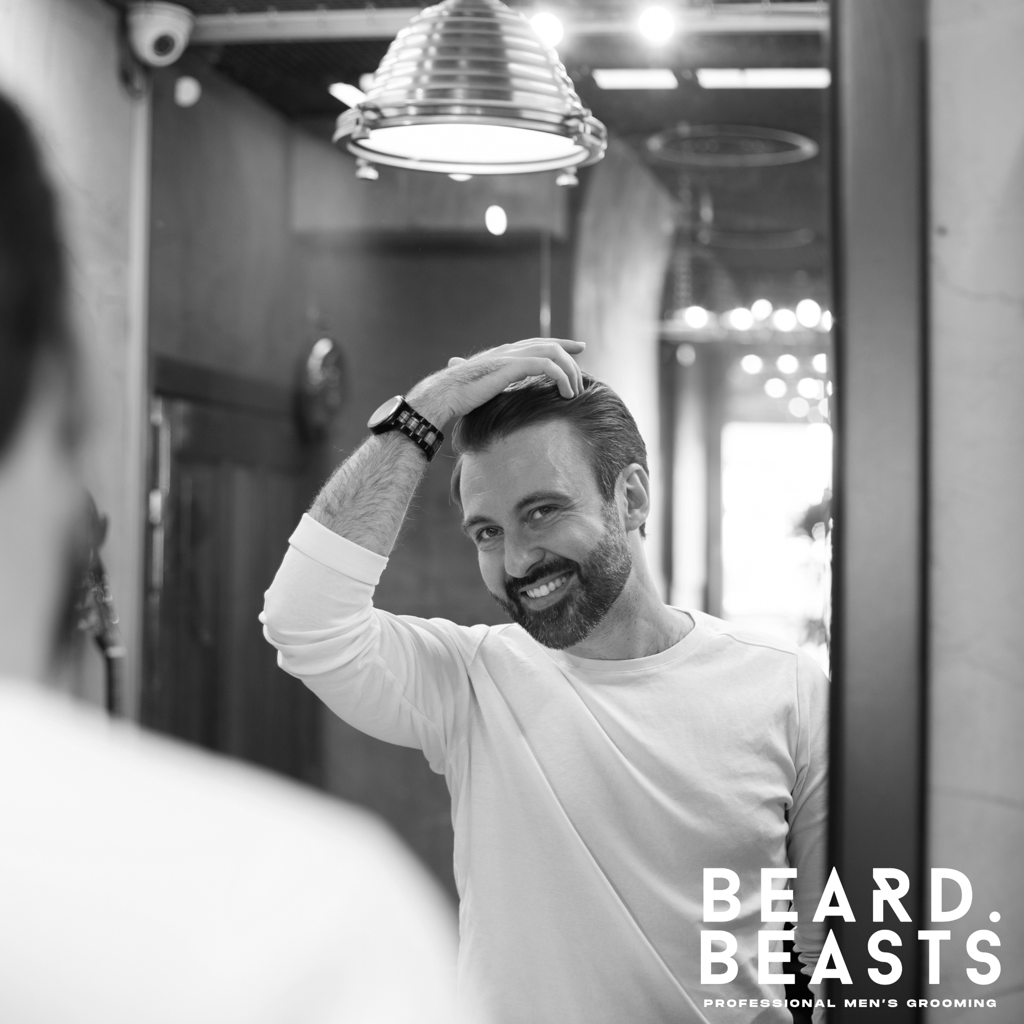 Smiling man styling his hair with matte clay in front of a mirror at a barbershop, wearing a white shirt.