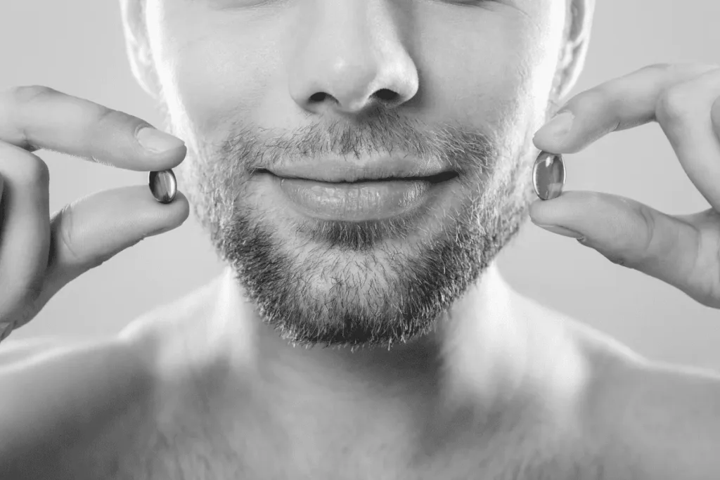 Man with a short groomed beard holding two different vitamin capsules to compare beard supplement options.