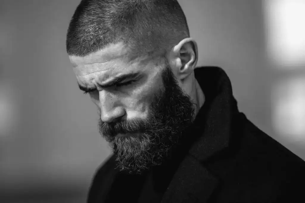 A dramatic black and white portrait of a man with a full, healthy beard and buzz cut, illustrating the clean, matte texture achieved by fixing a greasy beard.
