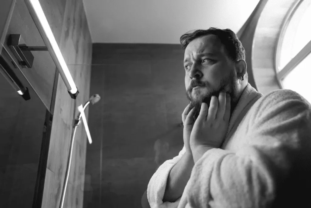 A man in a bathrobe closely examining his facial hair in the bathroom mirror to check for growth density and patches.