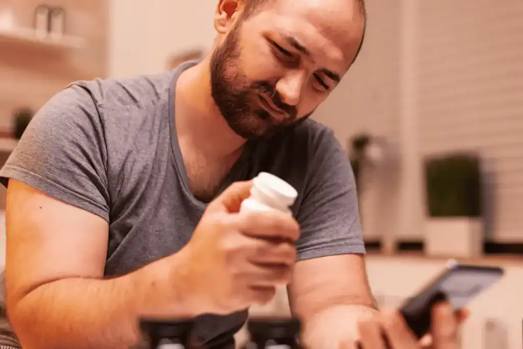 Man looking at beard growth supplement bottle while researching information on his phone