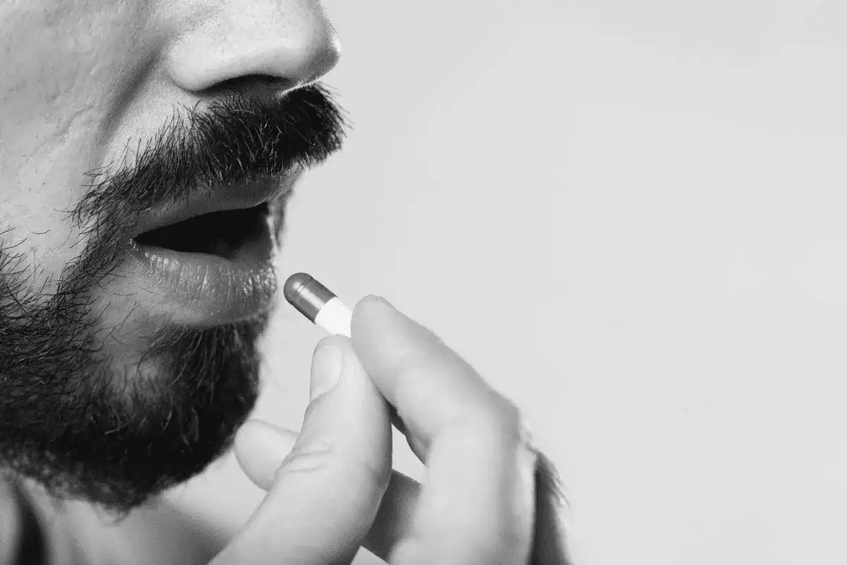 Close-up side profile of a bearded man placing a beard growth vitamin capsule into his mouth as part of his daily supplement routine.