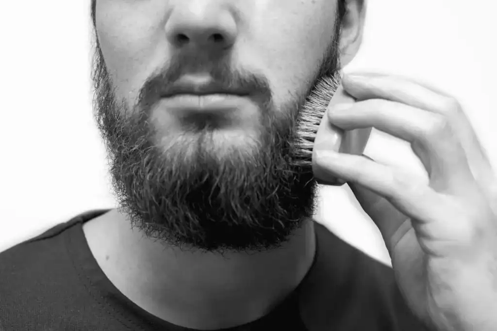 A close-up of a man using a wooden boar bristle brush to distribute natural oils and exfoliate the skin, a key step in fixing a greasy beard.