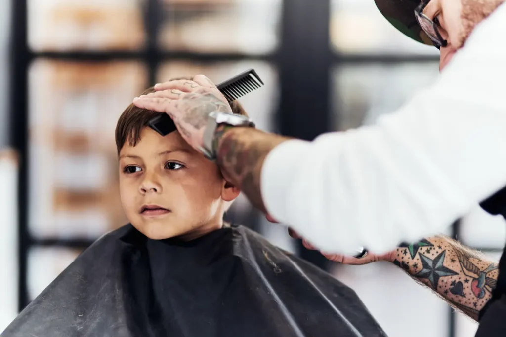 Young boy getting his hair cut at a barbershop while the barber uses a comb and scissors.