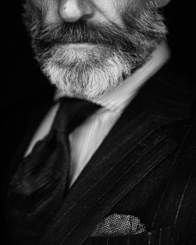 Close-up of a man with a full grey beard wearing a dark suit and tie