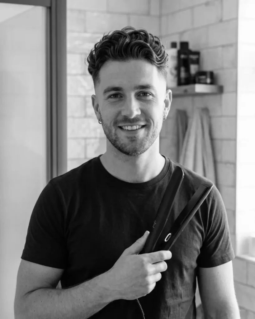 Man with short wavy hair holding a flat iron in a bathroom