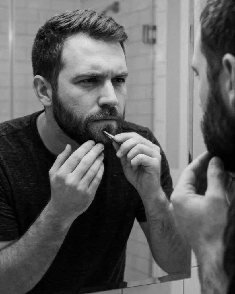 Man plucking white hairs from his beard in the mirror, showing why chasing individual grey beard hairs can make the beard look uneven.